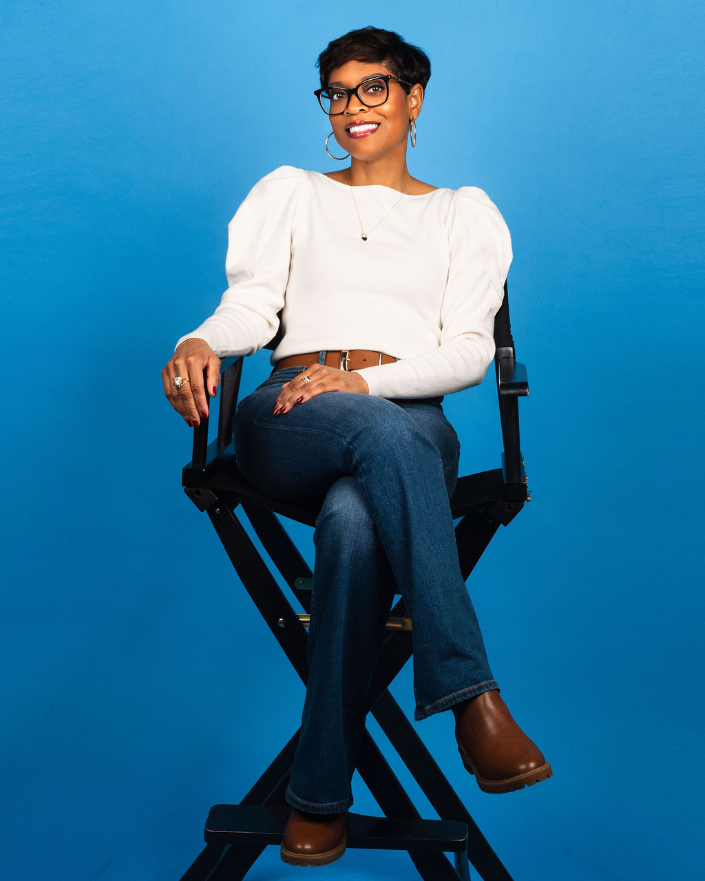 farrah sitting with her leg crossed in a director hair. in it, she is short hair, is smiling and wearing glasses
